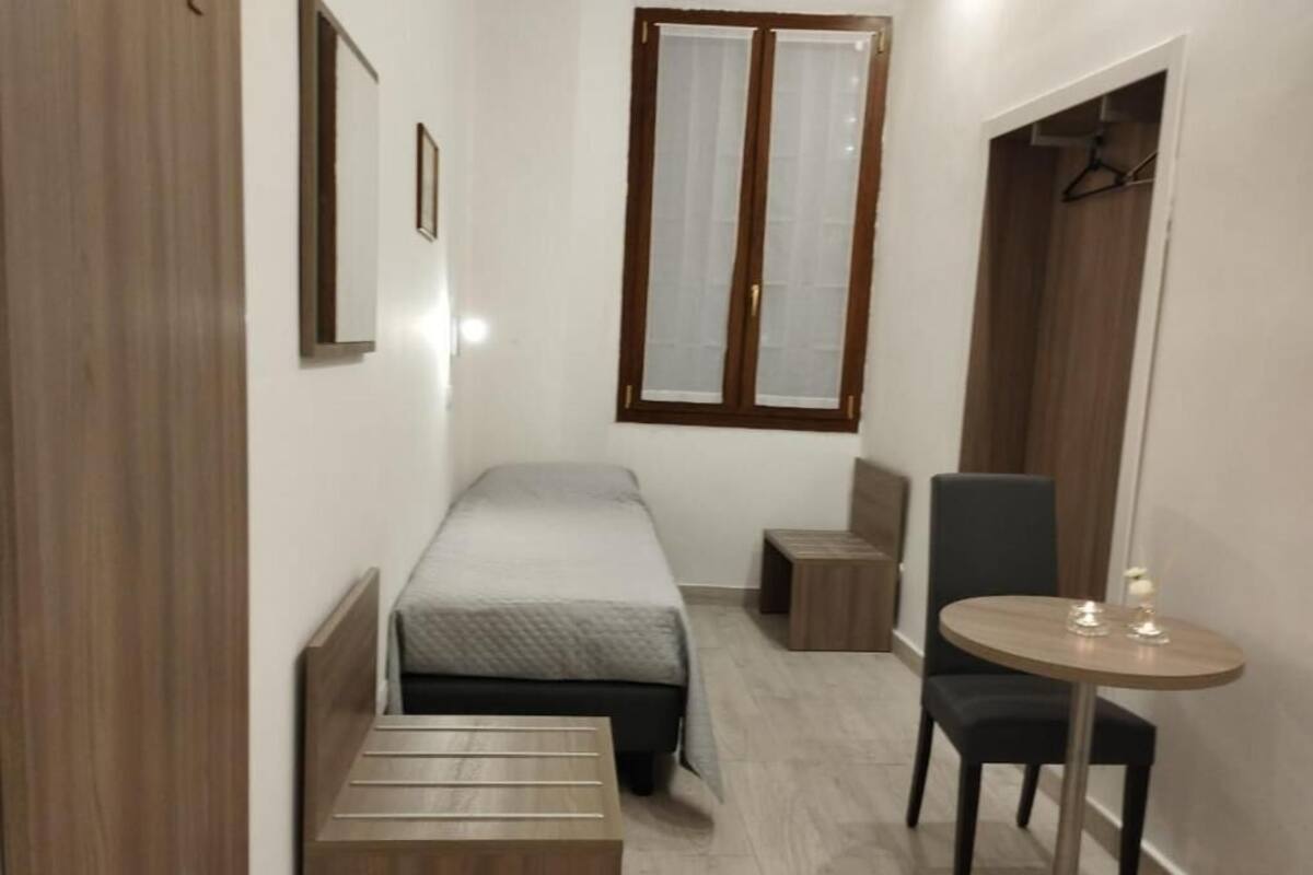 A small, compact room with a single bed, a small table with two chairs, a mirror, and a window with curtains.
