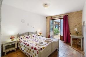 A bedroom with a bed, bedside tables, a window with curtains, a stone wall, and a view of the outside.