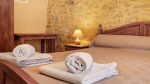 Una habitación de hotel con pared de piedra, una cama de madera con dos toallas blancas enrolladas, una mesita de noche de madera con una lámpara y un cuadro en la pared.