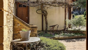 Un edificio de piedra con puerta de madera, una planta en maceta sobre una mesa y un carretón en el jardín.