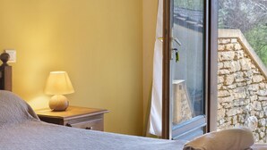 Un dormitorio con cama, mesita de noche con lámpara, y ventana con vista a un muro de piedra y árboles.