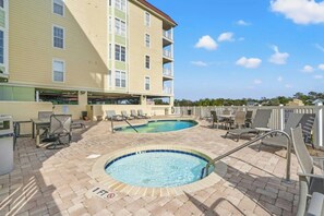 A rooftop pool area with two pools, lounge chairs, and a building in the background. - Ocean Marsh 404- 3 Bedroom in Windy Hill (North Myrtle Beach)