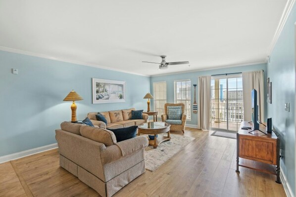 A spacious living room with a beige sofa, a wicker chair, a coffee table, and a TV stand. - Ocean Marsh 404- 3 Bedroom in Windy Hill (North Myrtle Beach)