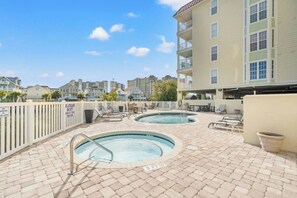 A rooftop pool area with two hot tubs and lounge chairs, surrounded by a white fence and a building with multiple balconies. - Ocean Marsh 404- 3 Bedroom in Windy Hill (North Myrtle Beach)