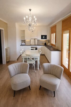 Une cuisine moderne avec des meubles blancs, un îlot central et un lustre. On y trouve deux chaises rembourrées devant une table à manger.