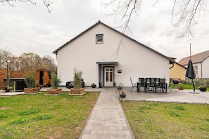A building with a paved entrance, a door, and a small window. There are outdoor tables and chairs, and a wooden barrel structure. - Holiday Home at Seeburgersee – Just 100 Meters from the Water (Seeburg)