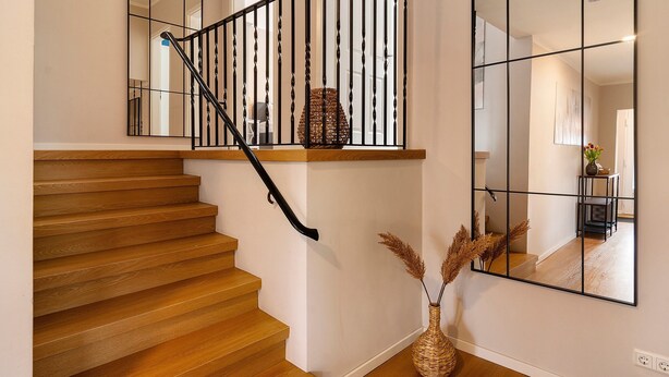 A wooden staircase with a black railing, a large mirror, and a vase with dried plants.