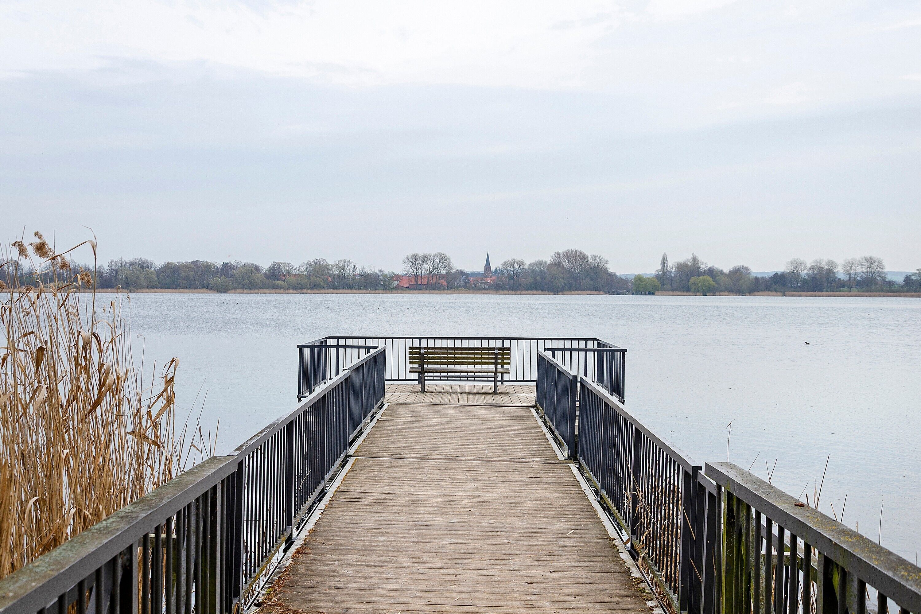 A wooden pier leading to a bench, with a calm water body and distant buildings.