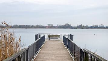 A wooden pier leading to a bench, with a calm water body and distant buildings.