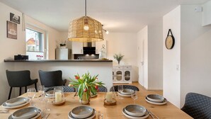 A dining area with a wooden table set for four, a hanging light fixture, and a kitchen in the background.