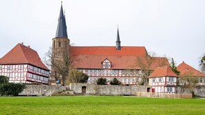 A historic building with a prominent tower and a red-tiled roof, surrounded by a stone wall and a green lawn.