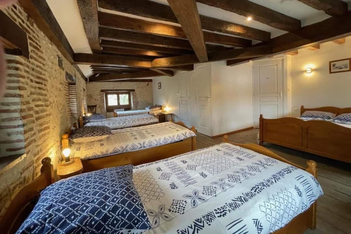 A rustic room with exposed wooden beams, two beds with patterned bedding, a brick wall, and a small window.