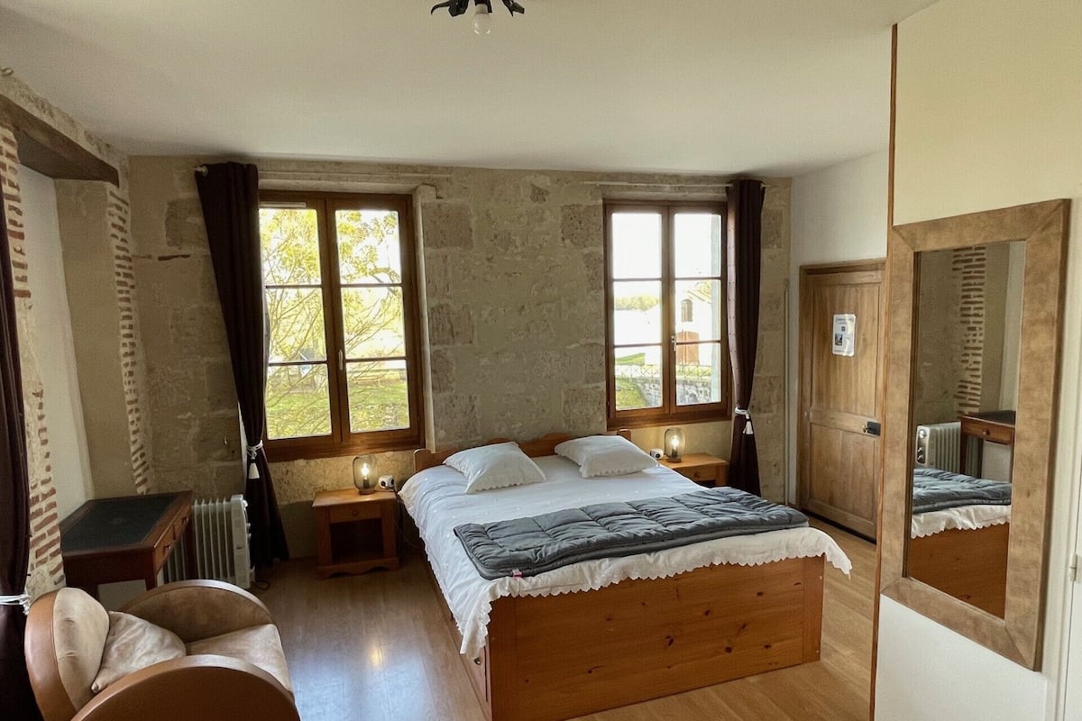 A bedroom with a wooden bed, two windows, a radiator, a chair, and a mirror.