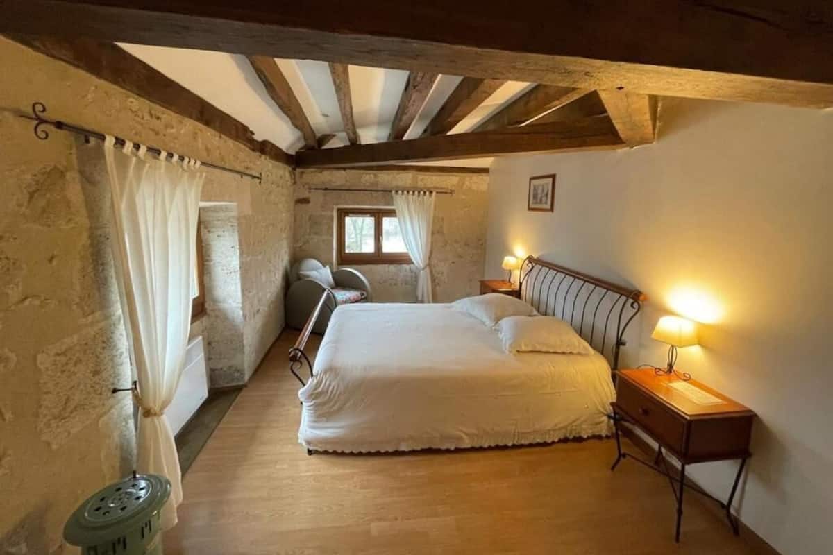 A bedroom with a large bed, wooden beams, a window with curtains, and bedside lamps.
