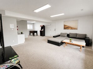 A modern living room with a sectional sofa, a coffee table, a foosball table, and a flat-screen TV. - Spacious Vernon Retreat w/ Game Room, BBQ Patio & Turf Yard (Vernon)