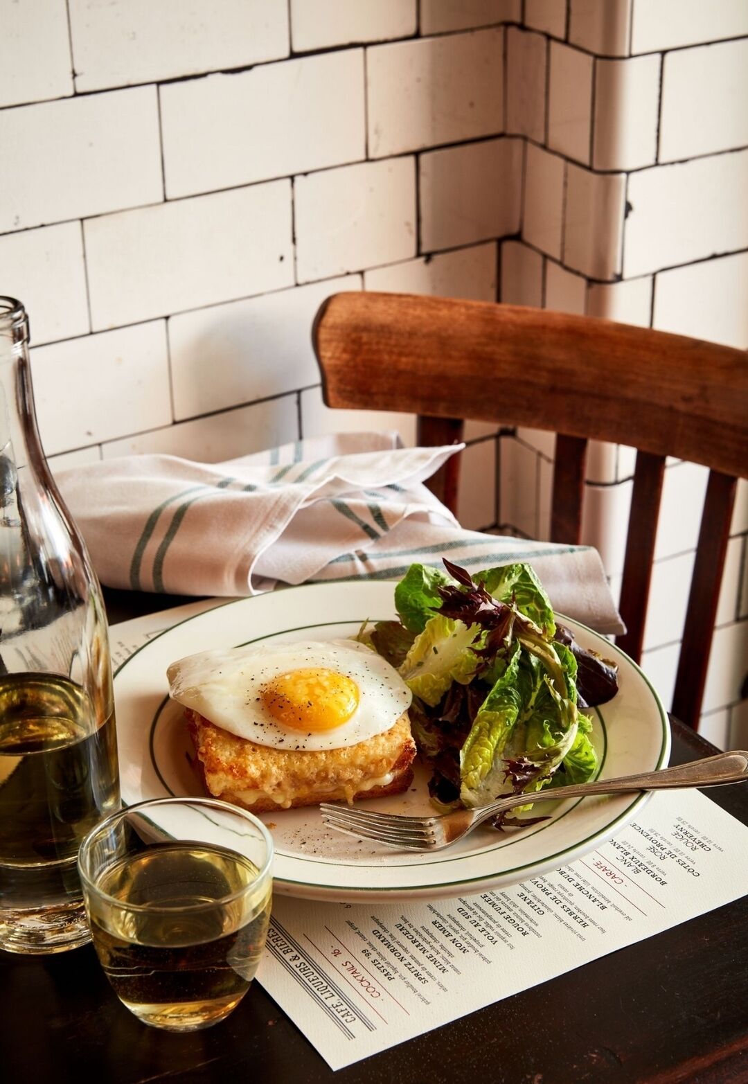 Une assiette avec un œuf au plat, une salade et une bouteille de vin, le tout posé sur une table avec un menu.