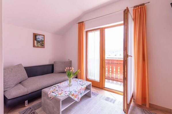 A room with a sofa, a table with a floral tablecloth, a vase with flowers, a painting on the wall, and a sliding glass door with curtains.