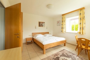A bedroom with a wooden bed, a nightstand, a chair, a window with curtains, and a door. - Holiday Apartment Isabelle Presseggersee (Presseggersee)