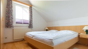 A single bed in a room with a wooden headboard, a window with curtains, a radiator, and a bedside table with a lamp.