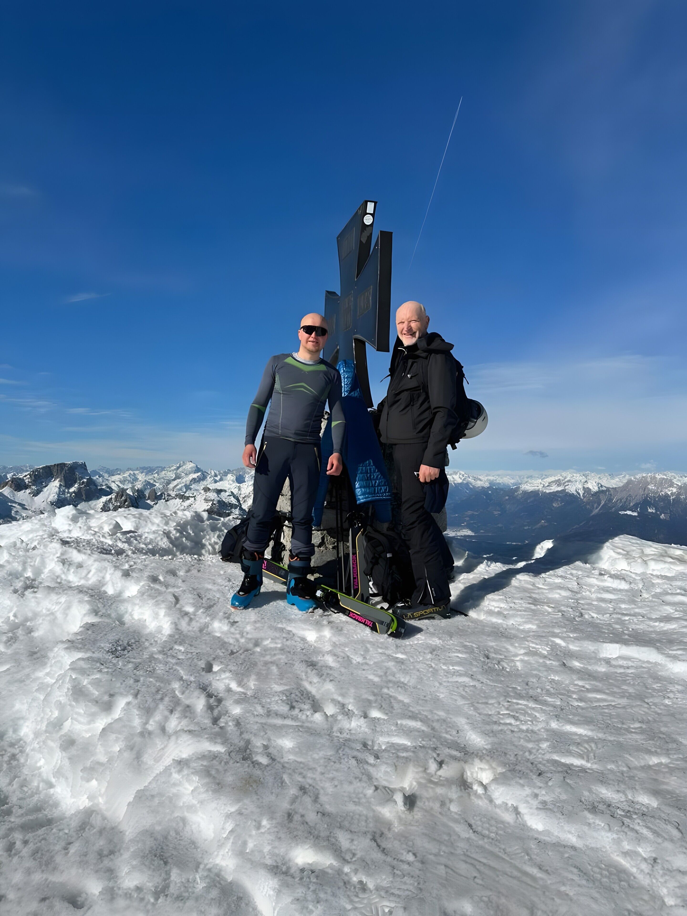 Two people standing on a snowy mountain with a clear sky, a signpost, and ski equipment.