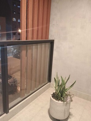 Ein Balkon mit Glasgeländer, einem Blumentopf und Blick auf ein Gebäude mit beleuchteten Fenstern.