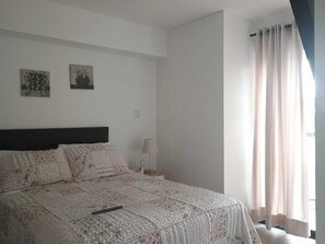 Ein Schlafzimmer mit einem Bett, einem Nachttisch, einer Lampe und einem Fenster mit Vorhängen.