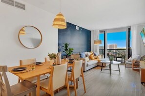 A modern living room with a dining area, a large round mirror, and a city view through large windows. - Bright Escape | 2BR Views | Resort | Gym & Pool (Hallandale Beach)