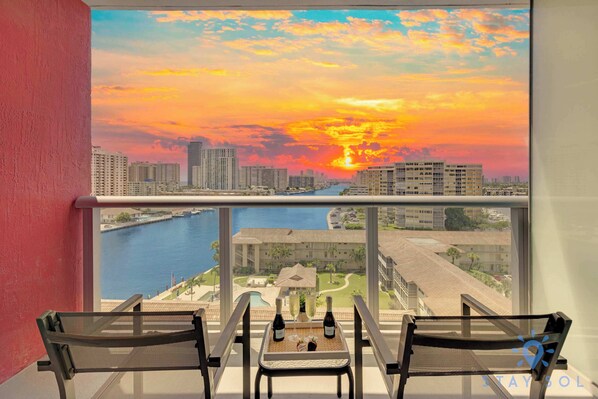 A balcony with a table set for two, overlooking a cityscape and a body of water during sunset. - Amazing Apartment, Pool and Gym.-Balcony View (Hallandale Beach)