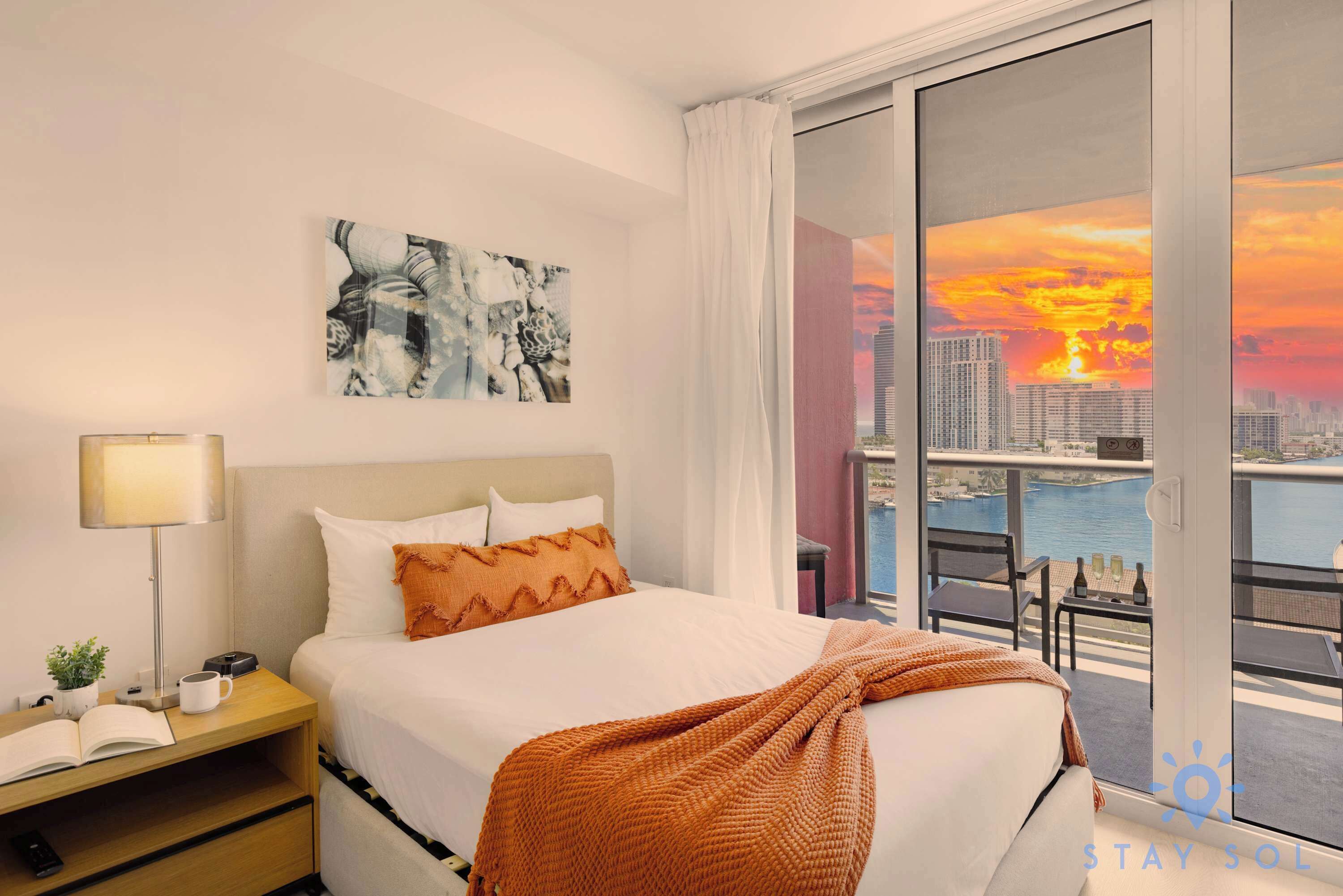 Camera d'albergo con un letto, un comodino, una lampada, un quadro e un balcone con vista sulla città.