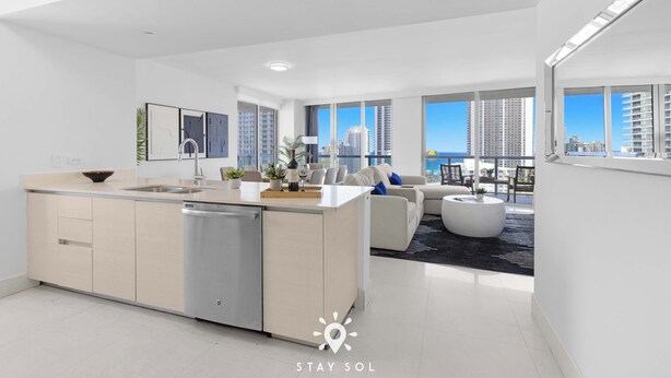 A modern apartment with a kitchen, dining area, and living room, featuring large windows and a view of high-rise buildings. - Beautiful Apt w/ Private Balcony + Incredible Pool (Hallandale Beach)