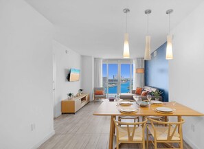 A modern living room with a city view, a dining area with a wooden table and chairs, and a cozy seating area with a sofa and cushions. - Cozy Getaway | 1BR + Gym | Beachside Vibe (Hallandale Beach)