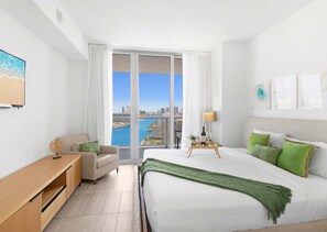 A modern hotel room with a large bed, a sofa, a desk, and a view of a cityscape and water. - Cozy Getaway | 1BR + Gym | Beachside Vibe (Hallandale Beach)