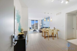 A modern, well-lit living area with a dining table, chairs, and a sofa. There is a large window providing a view of the city. - Stunning Views | Designed Luxe | Resort Stay (Hallandale Beach)