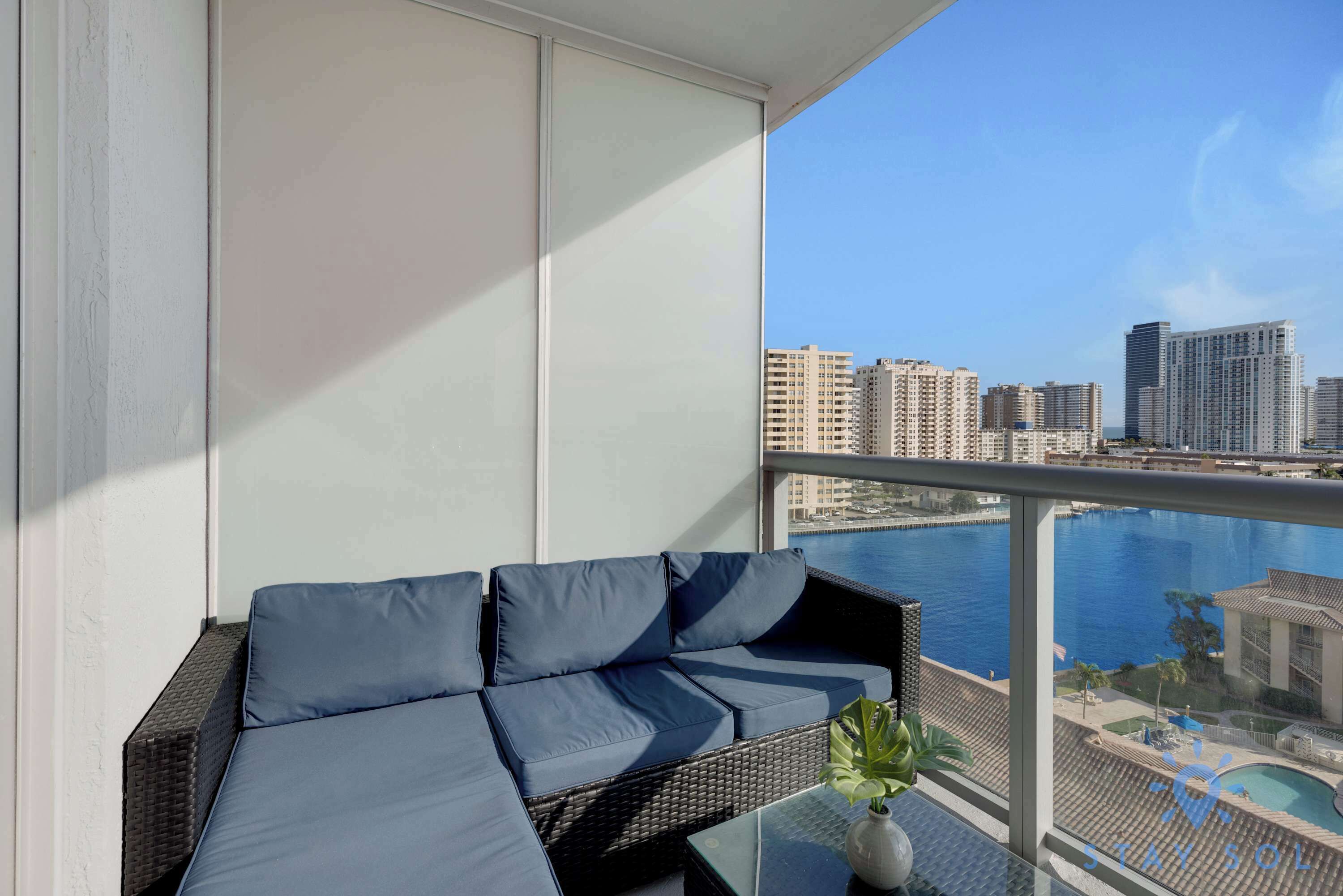 A balcony with a sofa and a small table, overlooking a cityscape and a body of water.