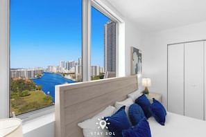 A hotel room with a large window offering a city view, a bed with blue and white pillows, and a bedside table with a lamp. - 3BR Apt w/ Private Balcony - Amazing Pool (Hallandale Beach)