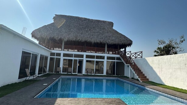 Una casa moderna con piscina, una struttura con tetto di paglia e una scala che conduce a un balcone.