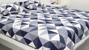 Un letto con copriletto a motivo geometrico in una stanza dalle pareti bianche e un comodino con una lampada.