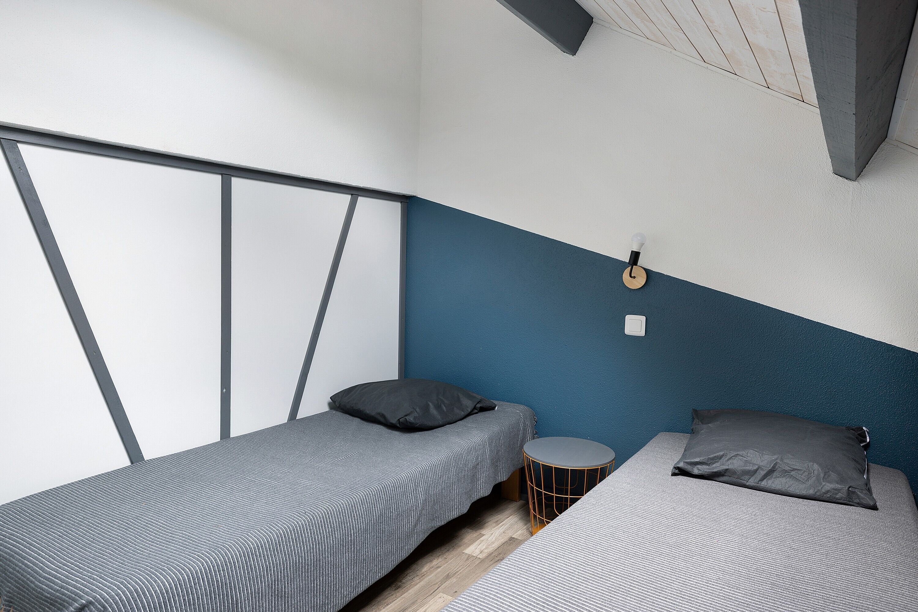 A small, modern room with two single beds, a round side table, and a view of the sky through a window.