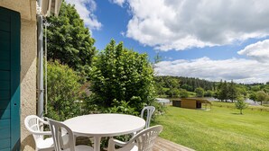 Eine Terrasse mit rundem Tisch und weißen Stühlen, ein Holzdeck und Blick auf ein Wiesenfeld und Bäume.