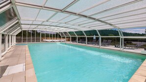 Ein überdachter Pool mit klaren Glaswänden, Liegestühlen und Blick auf eine bewaldete Landschaft.