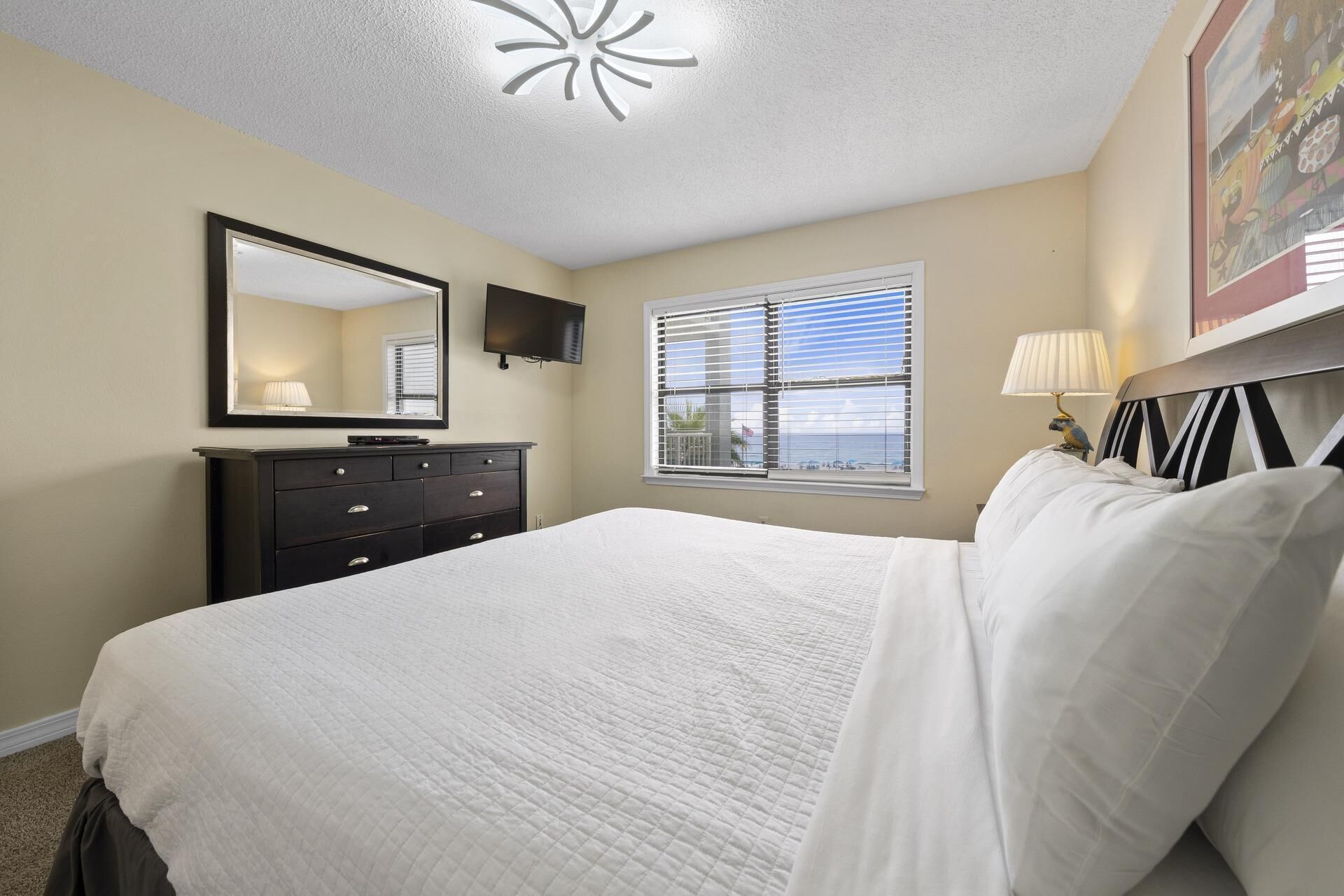A bedroom with a bed, dresser, TV, window with blinds, and a lamp.