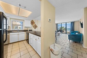 A kitchen with white cabinets and a black refrigerator, a dining area with a blue chair, and a view of the ocean through large windows. - Sunswept 201 (Orange Beach)