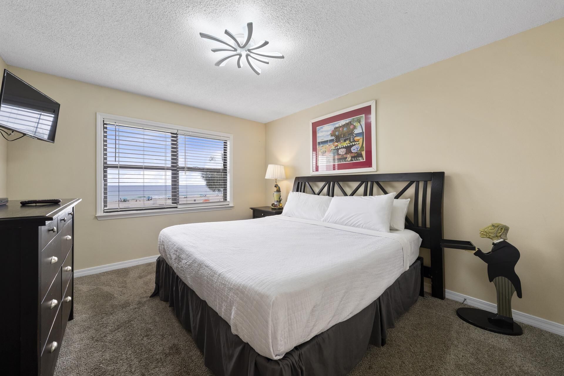 A hotel room with a bed, a desk, a lamp, a picture on the wall, and a ceiling light fixture.