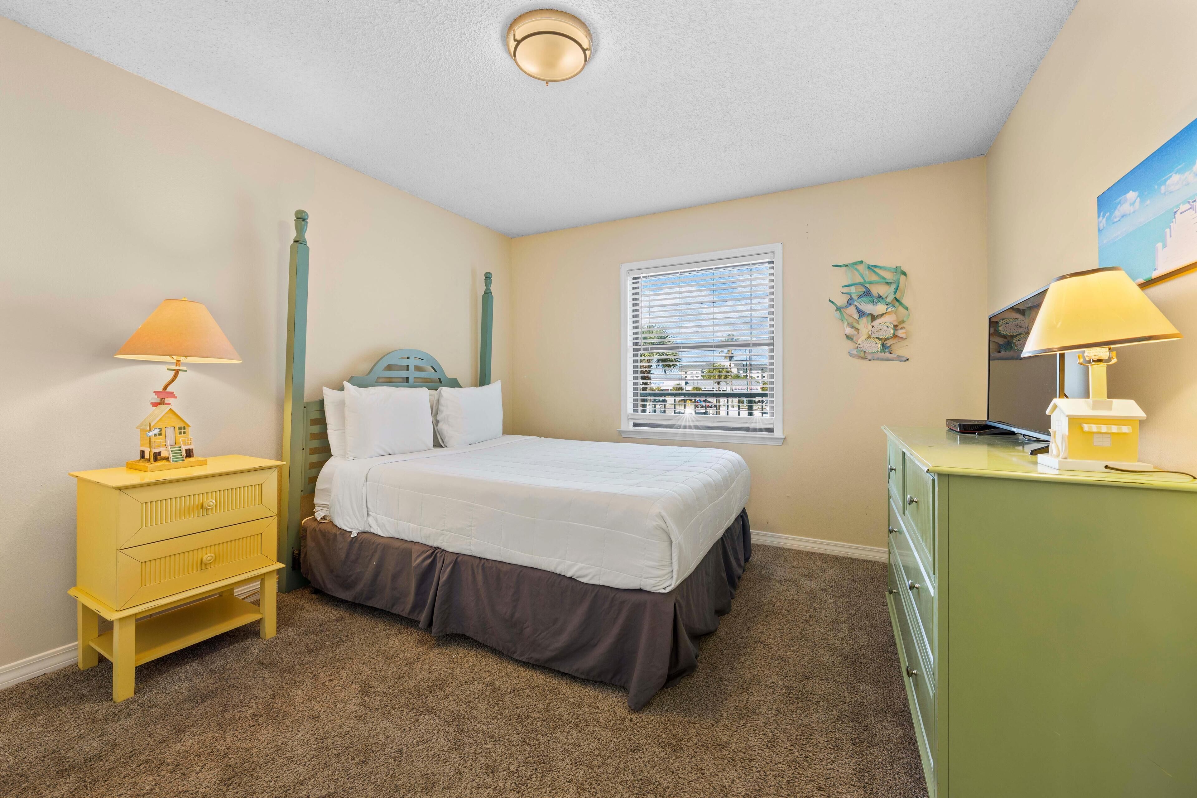 Une chambre à coucher comprenant un lit, deux tables de chevet, une commode, une fenêtre avec des stores et un luminaire au plafond.