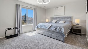 A bedroom with a bed, bedside lamps, a window with curtains, a ceiling fan, and a small bench. - Your Cozy Desert Retreat – Pool • Fire Pit • Convenient Location (Green Valley)