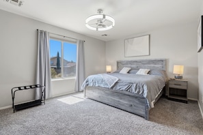 Une chambre à coucher avec un lit, des lampes de chevet, une fenêtre avec des rideaux, un ventilateur de plafond et un petit banc.