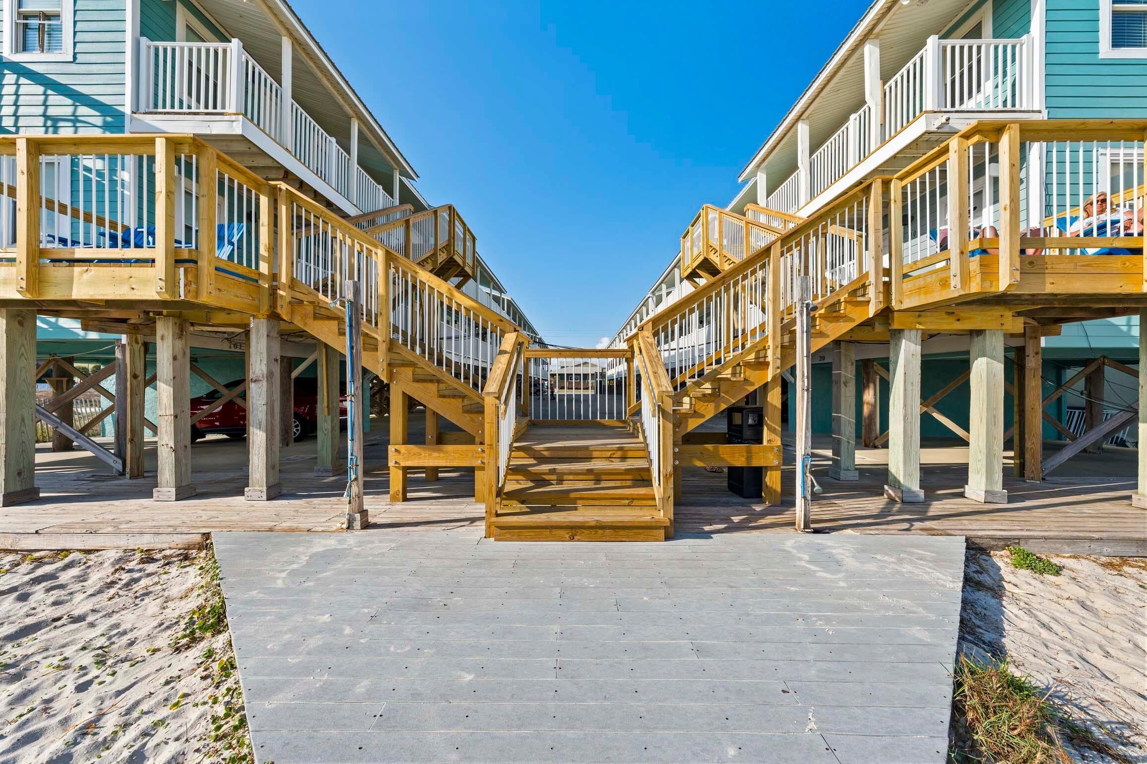 A wooden boardwalk leading to a beachfront property with multiple balconies and a clear sky.