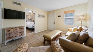 Un salon avec un canapé beige, un banc en bois, un téléviseur, une commode et une porte donnant sur une chambre.