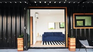Une porte ouverte donne sur un salon où l’on aperçoit un canapé bleu, des tables d’appoint en bois et un fauteuil noir.