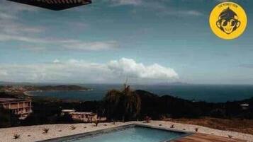 A pool with a wooden deck, a shower, and a view of the ocean and hills.